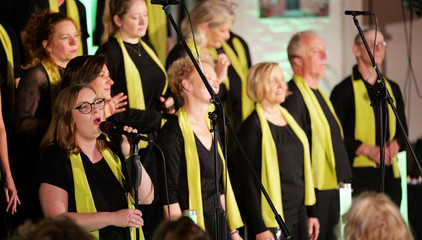 Mehrere Sängerinnen und Sänger stehen beim Singen mit grünen Schals vor Mikrofonen auf der Bühne beim Konzert des Albertinen Mitarbeitenden Gospelchors in der Kirche am Albertinen Krankenhaus in Hamburg.