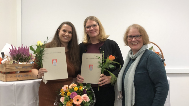 Drei Frauen stehen bei einer Abschlussfeier zusammen; zwei von ihnen halten Zertifikate und Blumensträuße nach bestandener Weiterbildung zur Hygienefachkraft.