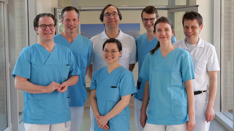 Das Team Des Mitralklappen-Zentrums des Albertinen Herz- und Gefäßzentrums am Standort Hamburg Schnelsen lächelt auf einem Flur in Arztkleidung in die Kamera