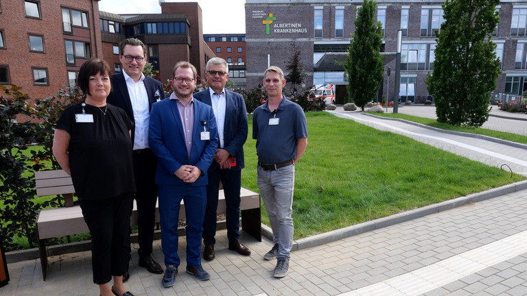 Ein Team mit dem Vorsitzenden der Konzerngeschäftsführung, Matthias Scheller, und Krankenhausgeschäftsführer Christian Rilz steht vor einer Ruhebank der neuen Zuwegung des Albertinen Krankenhauses in Hamburg-Schnelsen