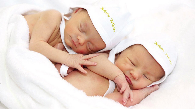 Zwei Neugeborene Babys liegen aneinandergekuschelt mkit weißenb Mützchen auf einer Decke