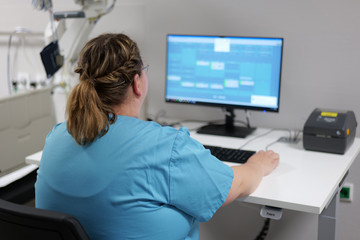 Mitarbeiterin in blauer Dienstkleidung sitzt mit dem Rücken zur Kamera an einem Computerarbeitsplatz im Behandlungsbereich.