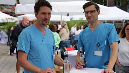 Zwei Ärzte in blauer Funktionsgleidung an einem Stehtisch beim Albertinen-Sommerfest für die Mitarbeitenden aus den unterschiedlichen Einrichtungen der Immanuel Albertinen Diakonie am Standort Schnelsen auf dem Platz vor der Kirche am Albertinen Krankenhaus.