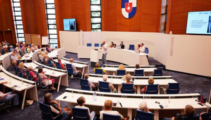 Blick über die besetzten Stuhlreihen auf die Bühne mit Stadtwappen und Monitoren im Sitzungssaal im Rathaus Norderstedt beim 1. Norderstedter Gelenktag für Betroffene und Interessierte zum Thema Arthrose. 