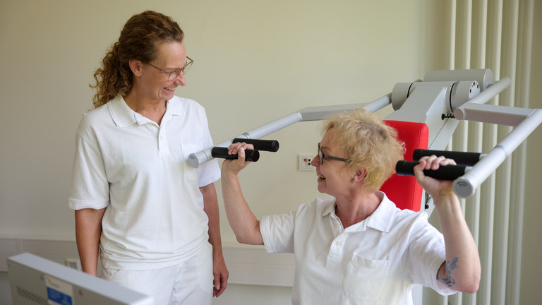 Eine Therapeutin sitzt an einem Trainingsgerät für Schulter- und Rückentraining und blickt zu einer Kollegin in weißer Arbeitskleidung im Interdisziplinären Therapiezentrum des Albertinen Krankenhauses in Hamburg