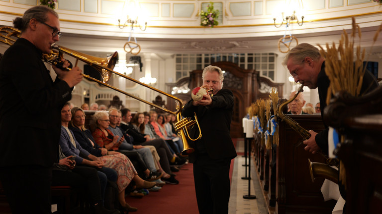 Ein Blechbläser läuft musizierend und in Begleitung von zwei anderen Bläsern des Trios "Windspiel" der NDR Bigband durch die mit Publikum besetzten Kirchengänge beim 10. Herzbrücke-Benefizkonzert im Hamburger „Michel“ der Albertinen-Stiftung.