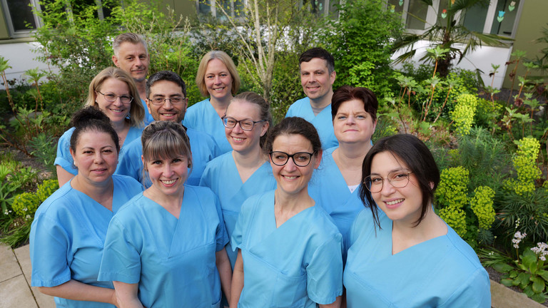 Mehrere Pflegekräfte aus dem Albertinen Zentrum für Altersmedizin strahlen in blauen Kasaks im Innenhof der Klinik in die Kamera